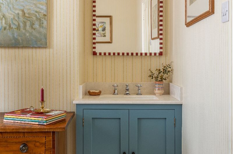 Compact bathroom with pretty wallpaper, blue vanity, wood storage and quirky wall decor.
