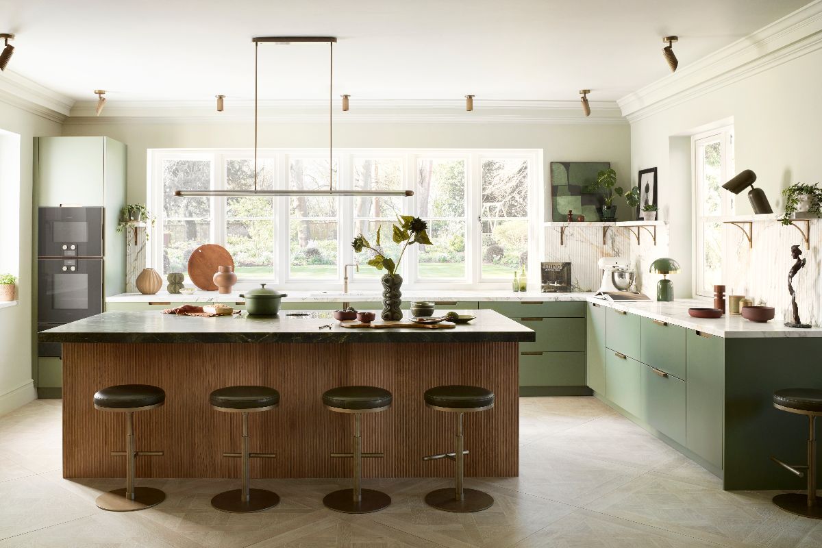 Open-plan kitchen with green cabinetry, a fluted wood island topped with rich green worktop, bar stools, and brass lighting.