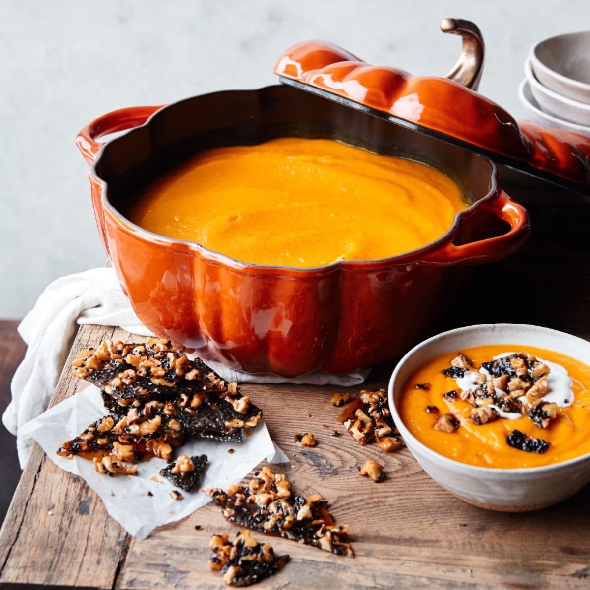 Pumpkin soup in a pumpkin-shaped dish with a small soup bowl next to it on a wooden chopping board.