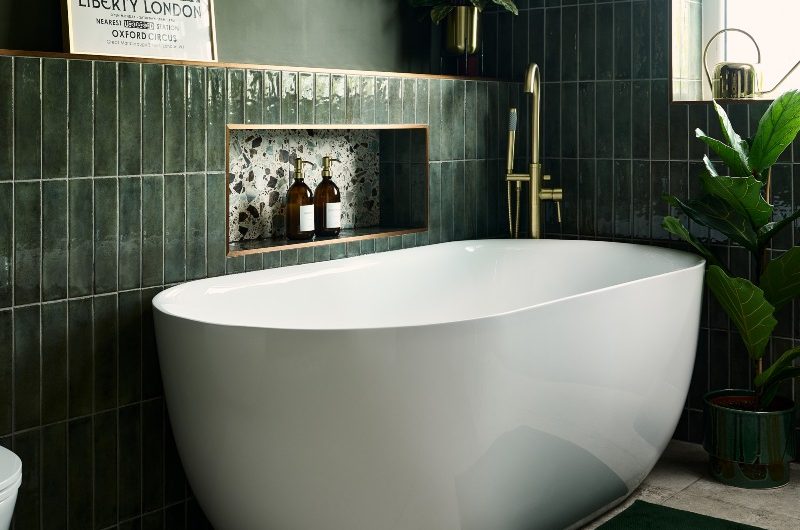 Green bathroom design with metro tiles, storage niche, white freestanding tub, gold tap, and a shelf for displaying decor.
