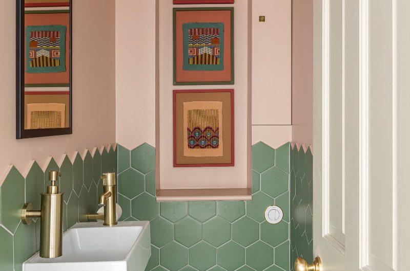 Cloakroom design with green hexagon tiles on the wall and floor, pink wall paint, a niche decorated with wall art, gold brassware and white sanitaryware.