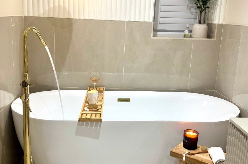 A neutral bathroom renovation with a freestanding tub, gold finishes, glossy wall tiles and reeded panelling.