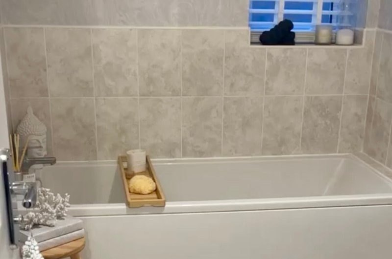 Before of a bathroom renovation with fitted tub, grey tiles and wood shelving.