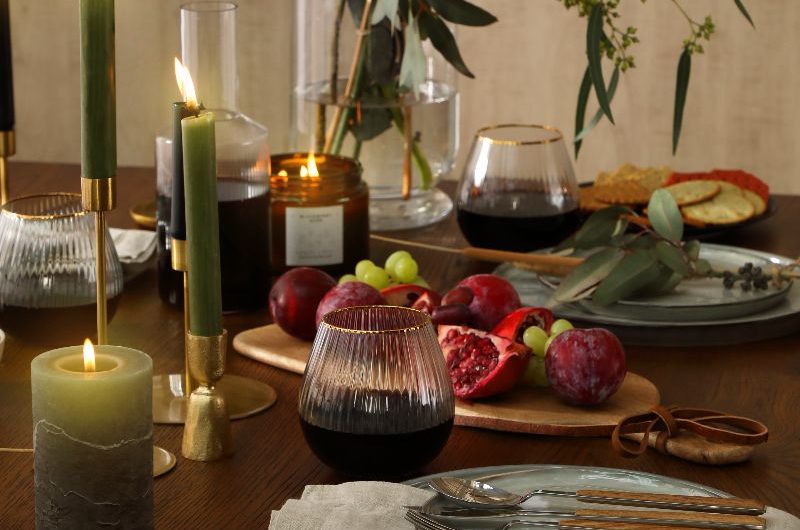 A closeup at a wood table decorated with sage green plates, fluted glasses, green candles, and a wood board with fruit.