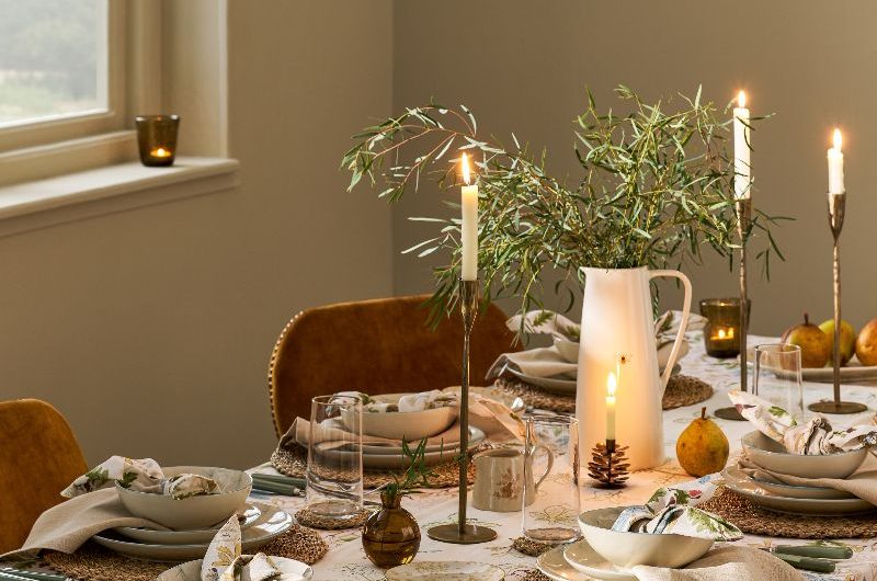 A dining table with forest-inspired tablecloth, napkins, brass candle holders, white ceramics, and velvet chairs.