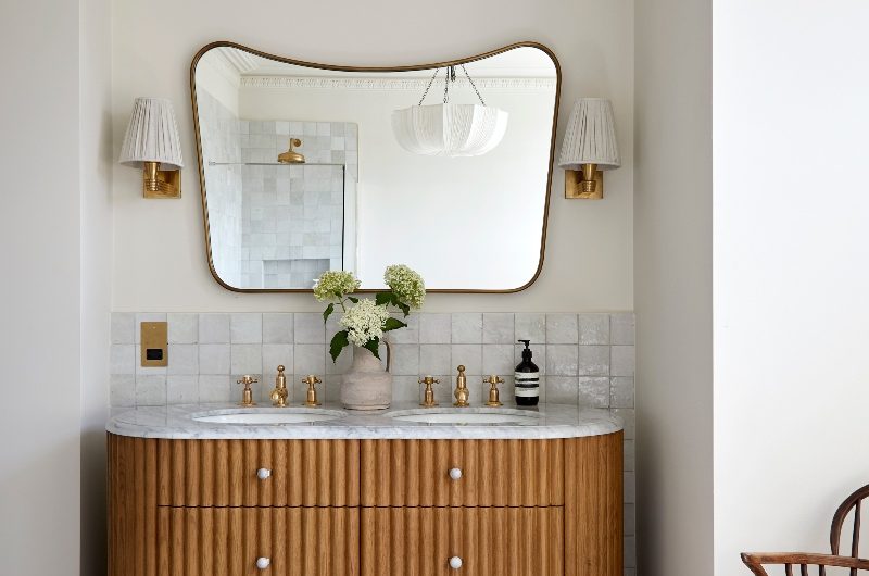A classic bathroom in a Victorian home with reeded wood vanity, zellige wall tiles, a curved mirror and wall lights.
