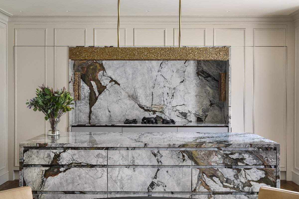 A glamorous scheme with panelled cabinetry, a statement hob zone with a single slab splashback, and an island clad in matching stone with a gold pendant above it.