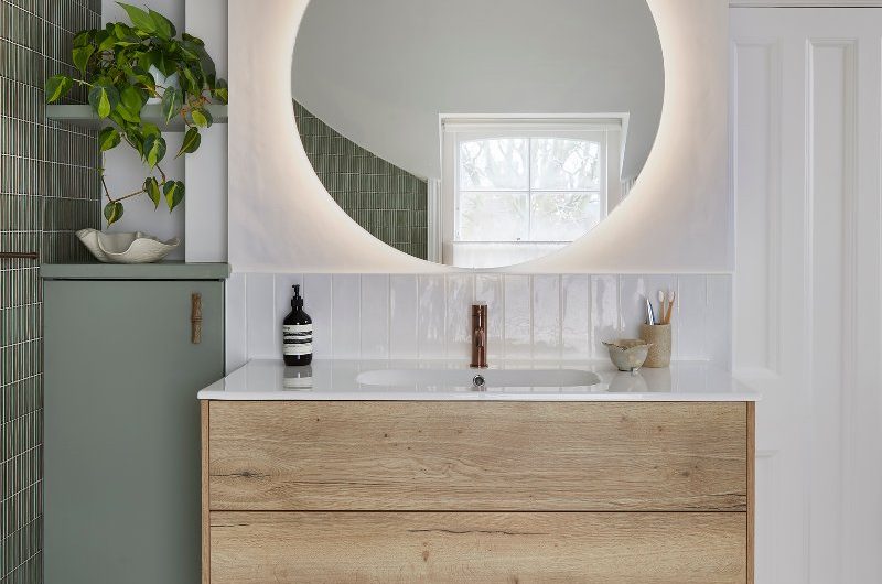 Green bathroom design with clever storage solutions, micro mosaic and white glazed metro tiles, and a vanity zone with a wood unit, inset sink, and circular smart mirror.