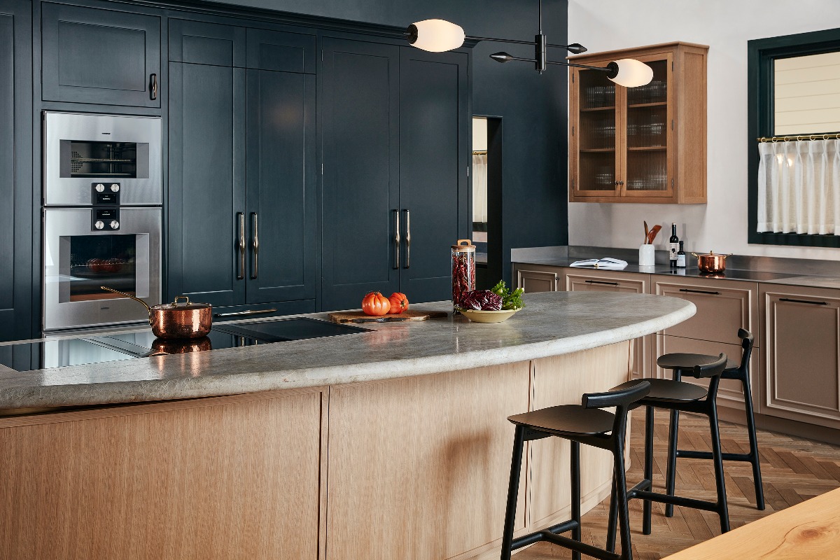 A classic scheme with a dark blue run of cabinetry, neutral base units and a curved island with a tapered base, and a bullnose countertop in leathered quartzite.