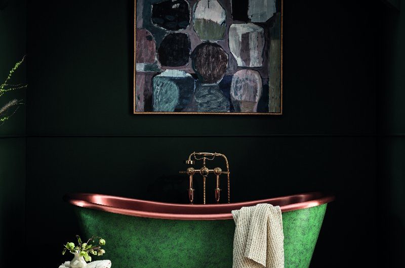 A dark green bathroom design with a green and copper freestanding bath, classic bath filler, wall art and wood flooring.