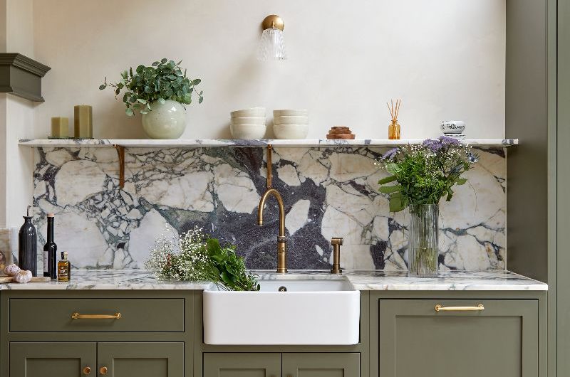 The sink zone of the dark green kitchen features a butler sink, brass tap and matching handles, a stone open shelf, and plaster on the walls.
