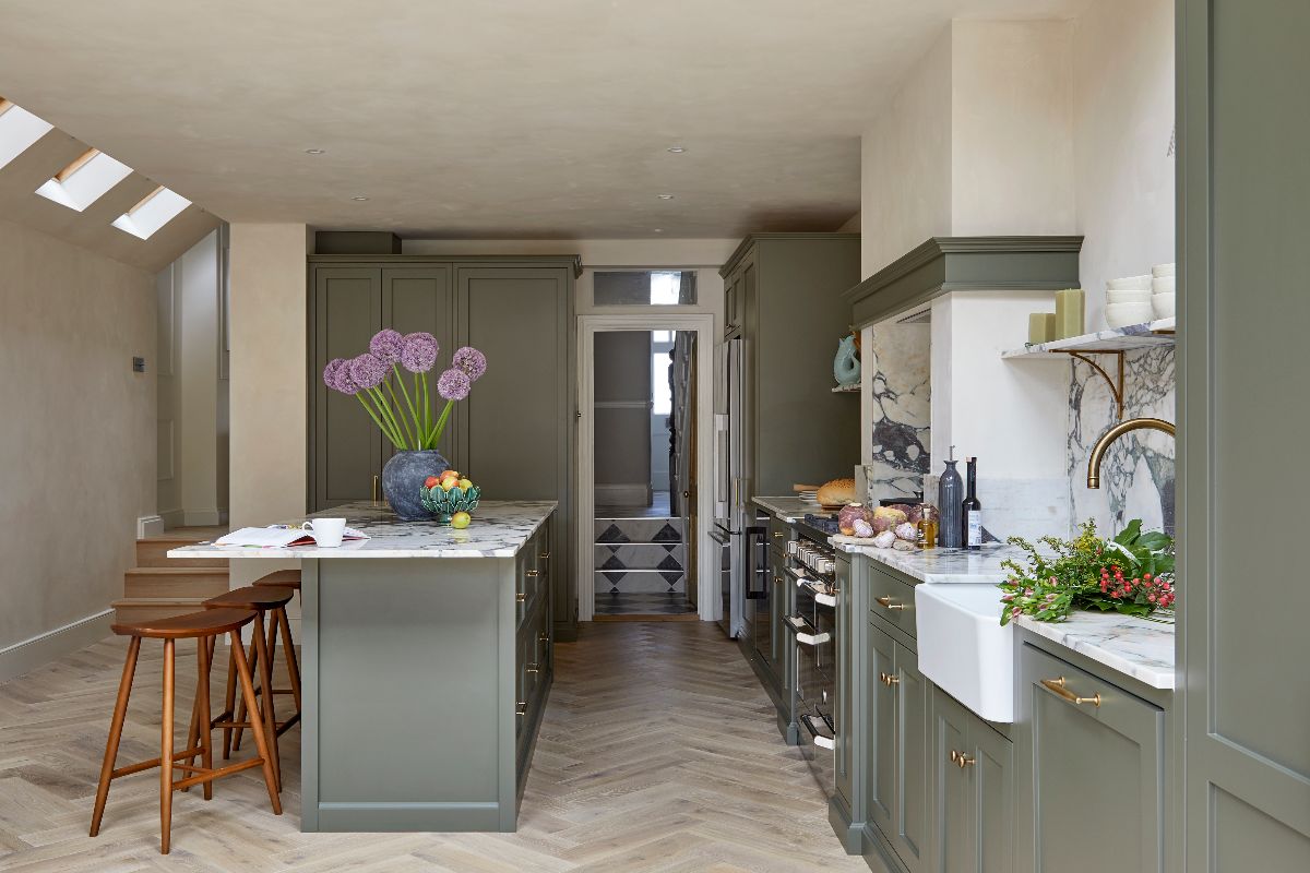 A dark green kitchen design in an L-shape with a central island, herringbone planks on the floor, plaster walls, and statement marble.