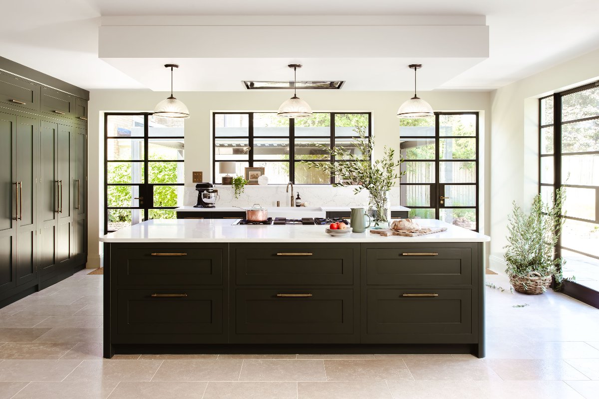 Bright scheme flooded with light through steel-framed glazing, dark green cabinetry, stone flooring and white worktops.