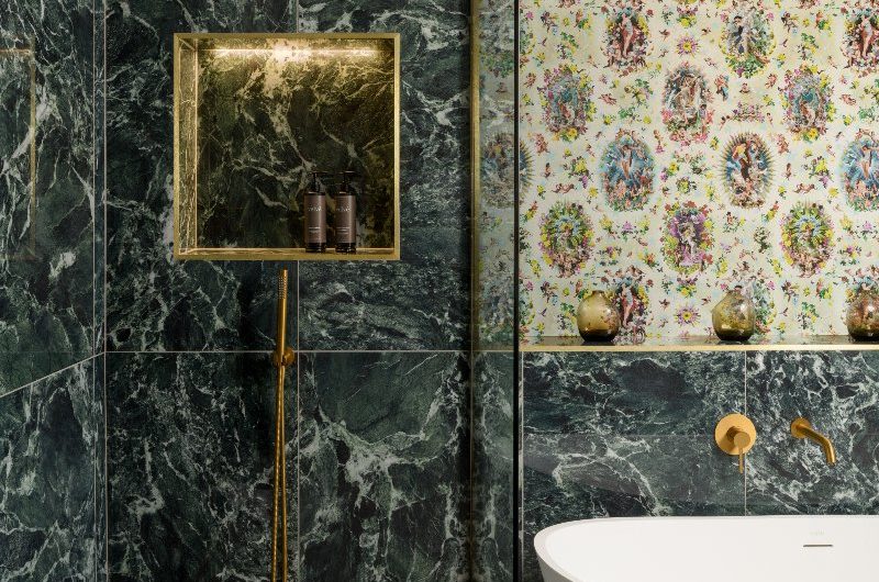 A statement bathroom design with painted ceiling, patterned wallpaper, and a green tile clad walk-in shower with an illuminated storage niche.