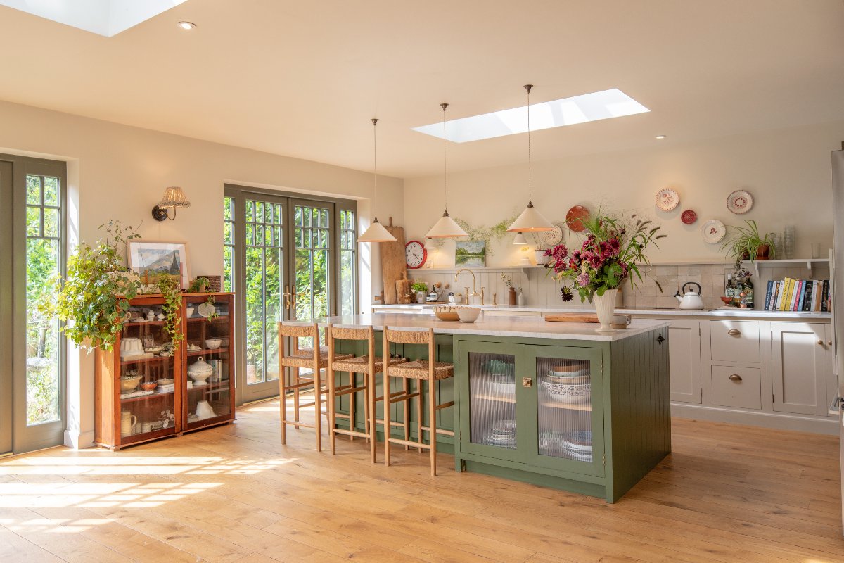 A bright kitchen design with cream units, zellige tiles, wood flooring, and a green island with fluted display cabinet and seating.