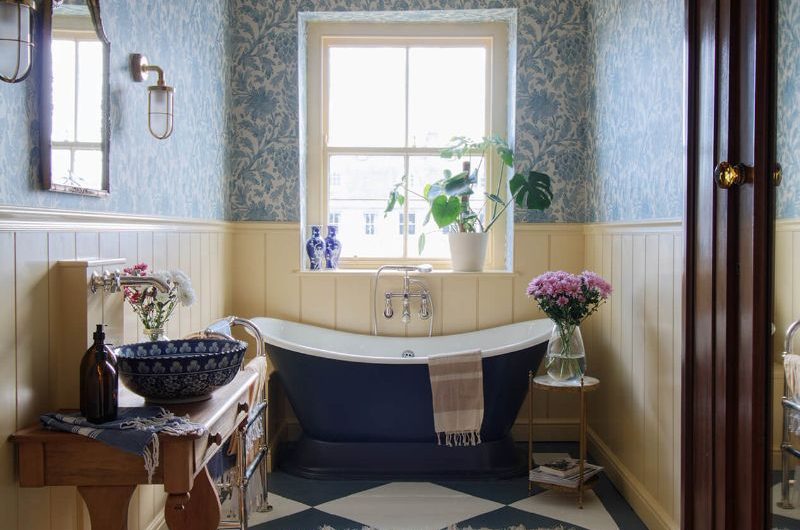 A classic bathroom design with yellow wall paint, floral wallpaper, checkered flooring and a rustic rug, a blue tub and wood vanity unit.