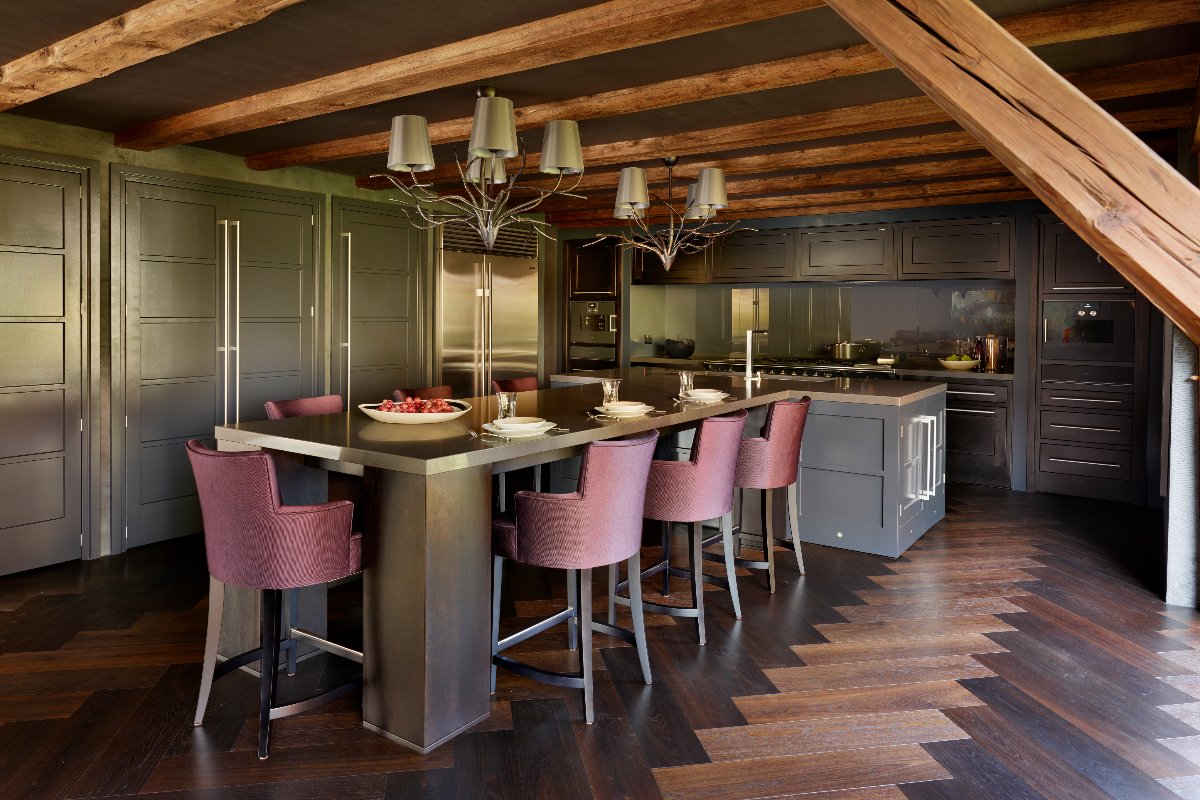 A statement kitchen design with a central island with an extra-long dining table style extension, luxurious burgundy chairs, herringbone flooring, and exposed wood beams.