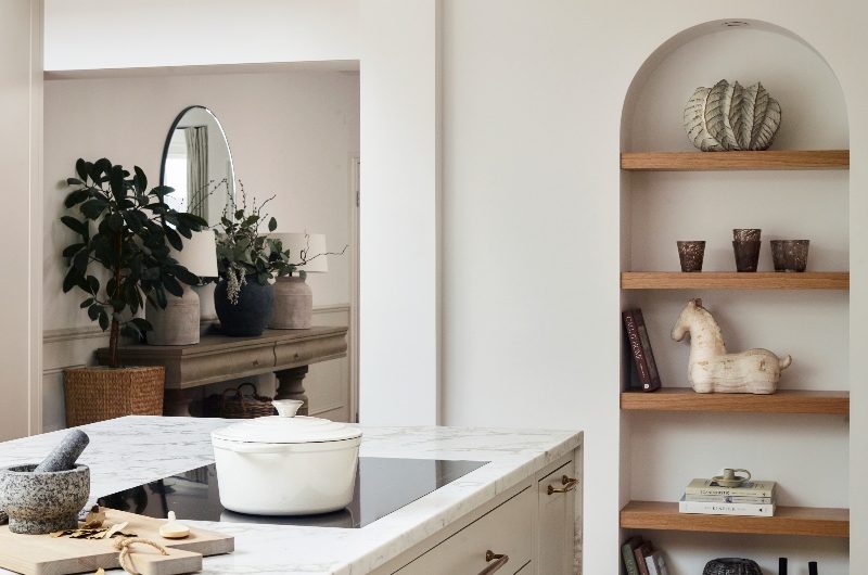A closeup at the kitchen island with neutral base and topped with veiny surfaces, with a built-in hob and an adjacent display area on the opposite wall.