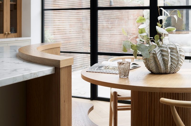 A dining zone with bespoke seating, fluted table, and wood chairs, overlooking the garden through steel-framed doors.