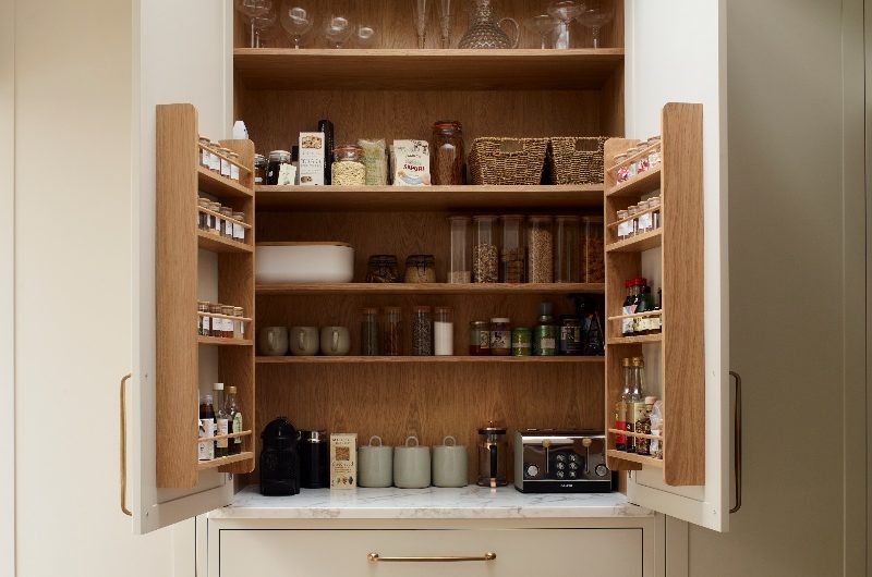 An open breakfast cupboard in a neutral kitchen with sleek cabinetry, gold hardware, oak interiors, and stone surfaces.
