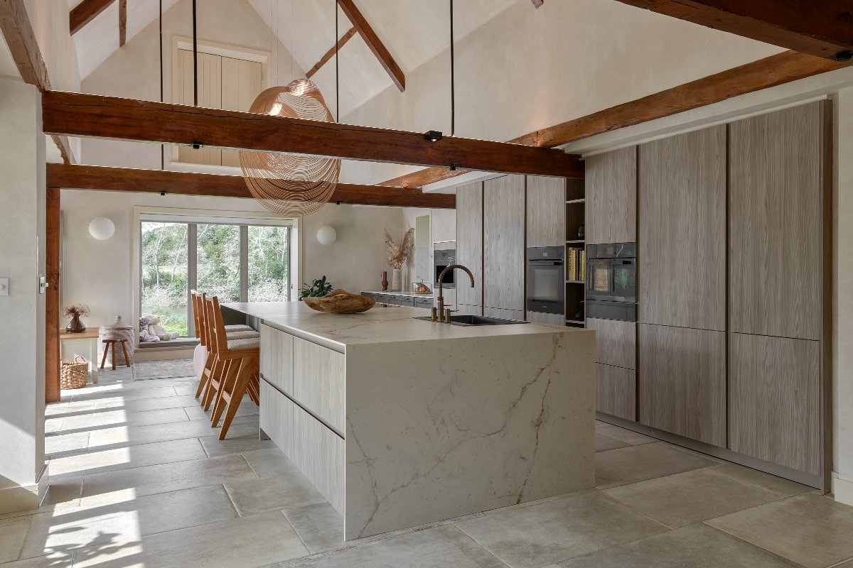 An open-plan scheme with exposed beams, a run of wood cabinetry with built-in appliances and a stone-clad island with shelves, sink and tap, and breakfast bar with seating.