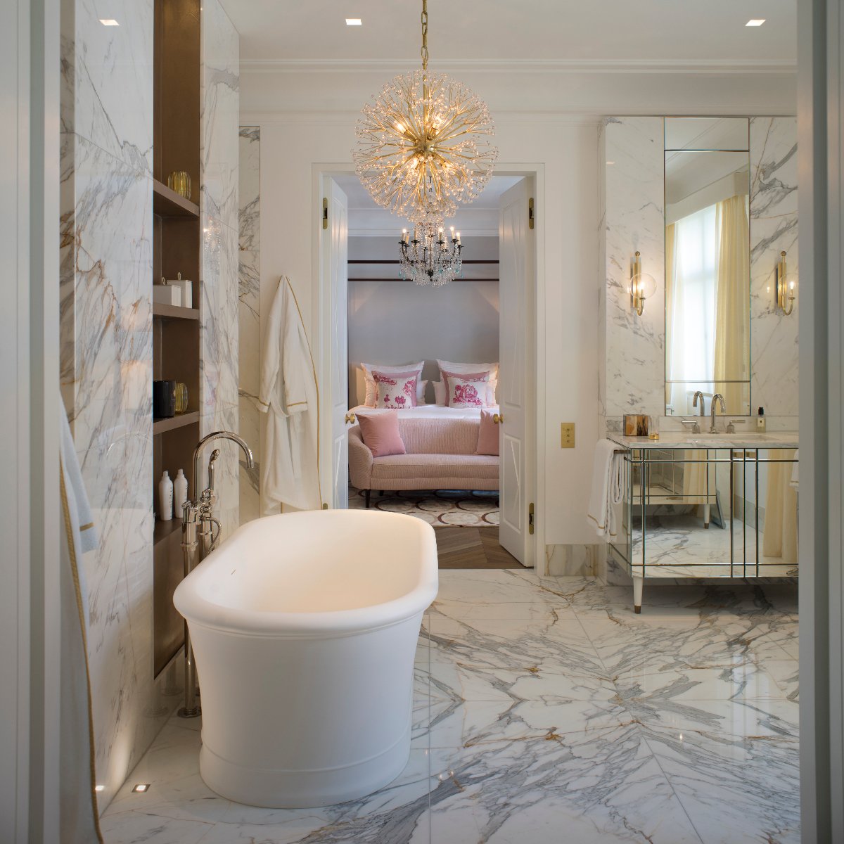 Luxury bathroom with marbled surfaces, a mirrored vanity, statement lighting, and a freestanding tub.