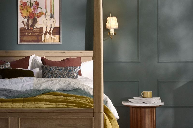 A boutique hotel bedroom with a four poster wood bed, blue wall paint, a reeded side table, and a patterned rug zoning the area.