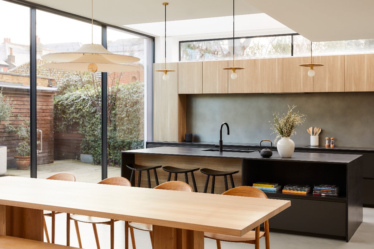 Tall glazing flooding an open-plan room with light with wood and black cabinetry, a central unit, with sink and tap, storage and seating, and a wood dining zone next to it.