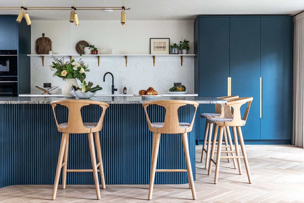 A bright scheme with tall cabinetry and a sink zone on one wall, a fluted island with coloured stone worktop, oak bar stools, and a gold pendant light.