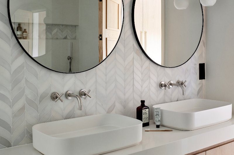 A double vanity space with a wood handleless unit, countertop basins, and circular black mirrors with marble mosaic tiles behind.