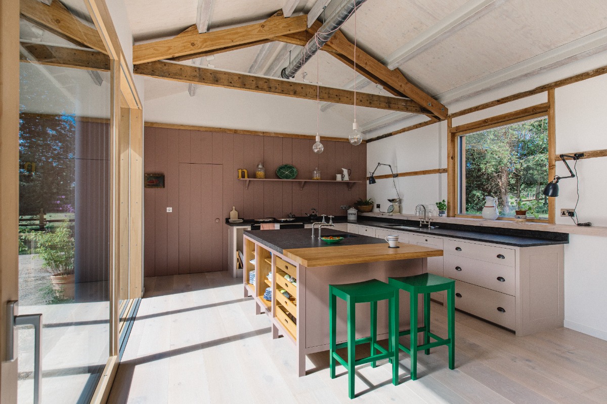 Dusty pink cabinetry topped with dark worktops, with a central unit with open storage, matching dark worktop with a wood breakfast bar and bright green stools.