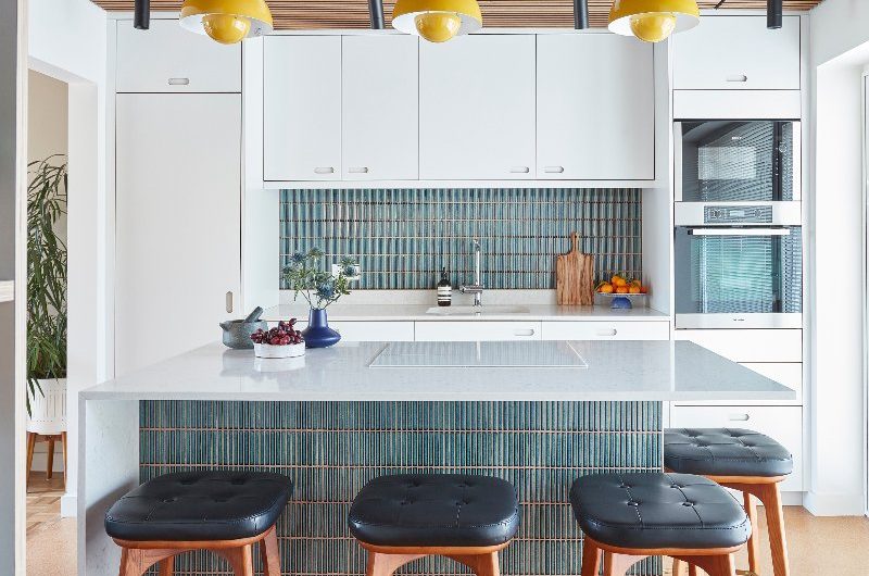 A modern white kitchen with built-in appliances, a compact island clad in green kitkat tiles with a waterfall edge and seating.