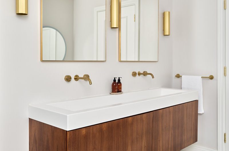 Vanity zone with twin mirrors and lights, and  a wall-mounted wood unit with white countertop surface and matching inset sink, and gold hardware.