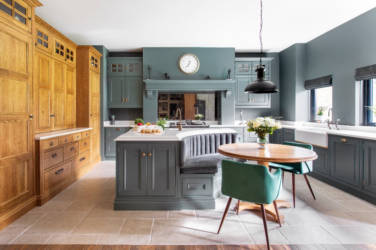 A classic scheme with dark green cabinetry, a wall of wood units, and a central multi-functional hub with a sink and tap, curved seat, wood dining table and two green chairs.