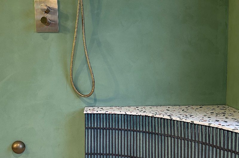 A wetroom style bathroom with green microcement, a shower seat, kitkat tiles, and terrazzo flooring.

