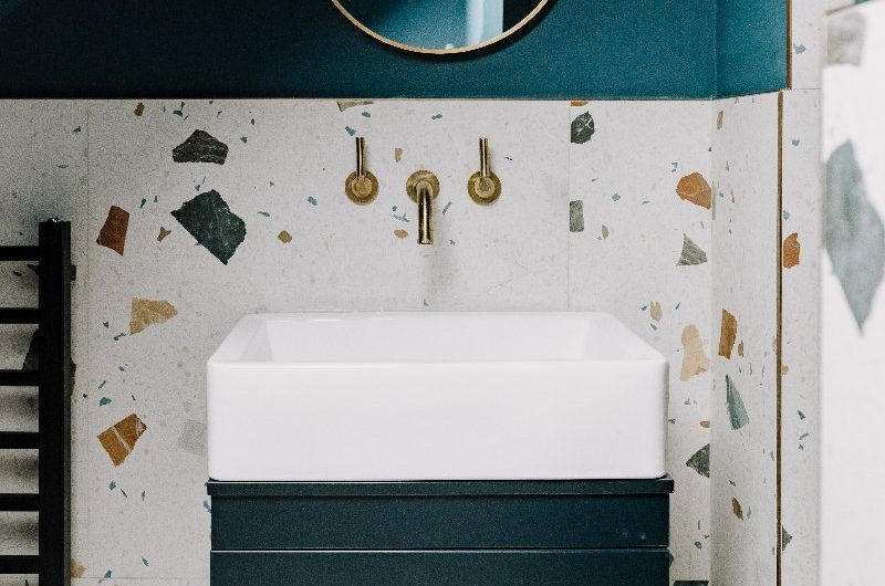 A modern design with large-format patterned tiles, a blue vanity unit with a white basin, gold brassware, and a circular mirror.