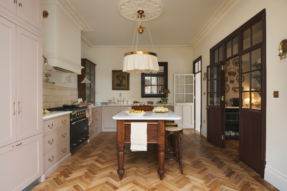 An L-shaped kitchen painted in dusty pink, with wood flooring, and a freestanding wood island with white worktop and seating.