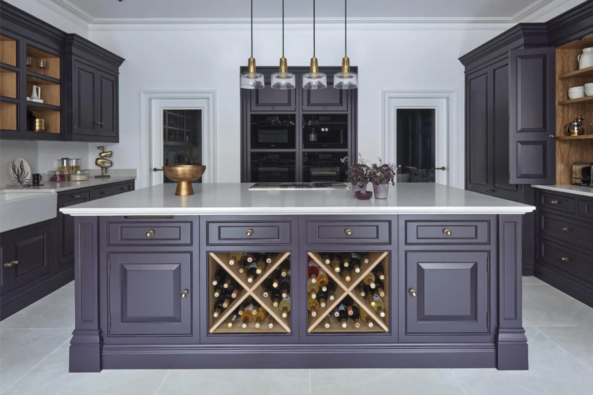 An island finished in a dark purple shade, with bottle storage, a creamy white worktop, natural oak accents, and brass hardware.