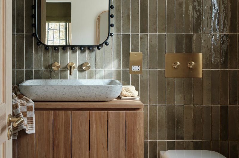 A green bathroom design with olive metro tiles, wood vanity unit, speckled basin, terrazzo flooring, curved toilet, and bobbin mirror.