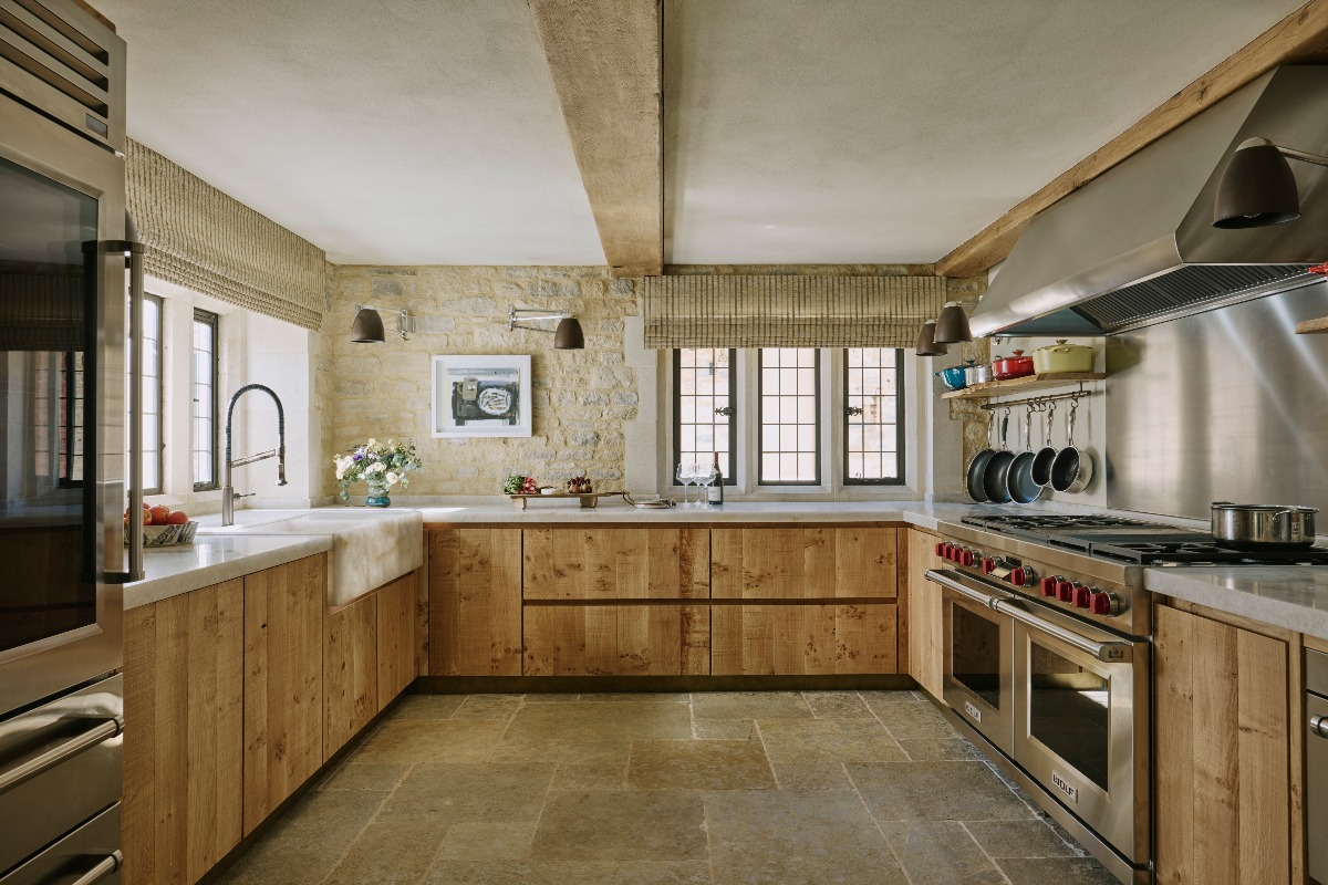 A U-shaped kitchen design with stainless steel appliances, wood cabinets, stone flooring, exposed brick on the walls and wood beams.