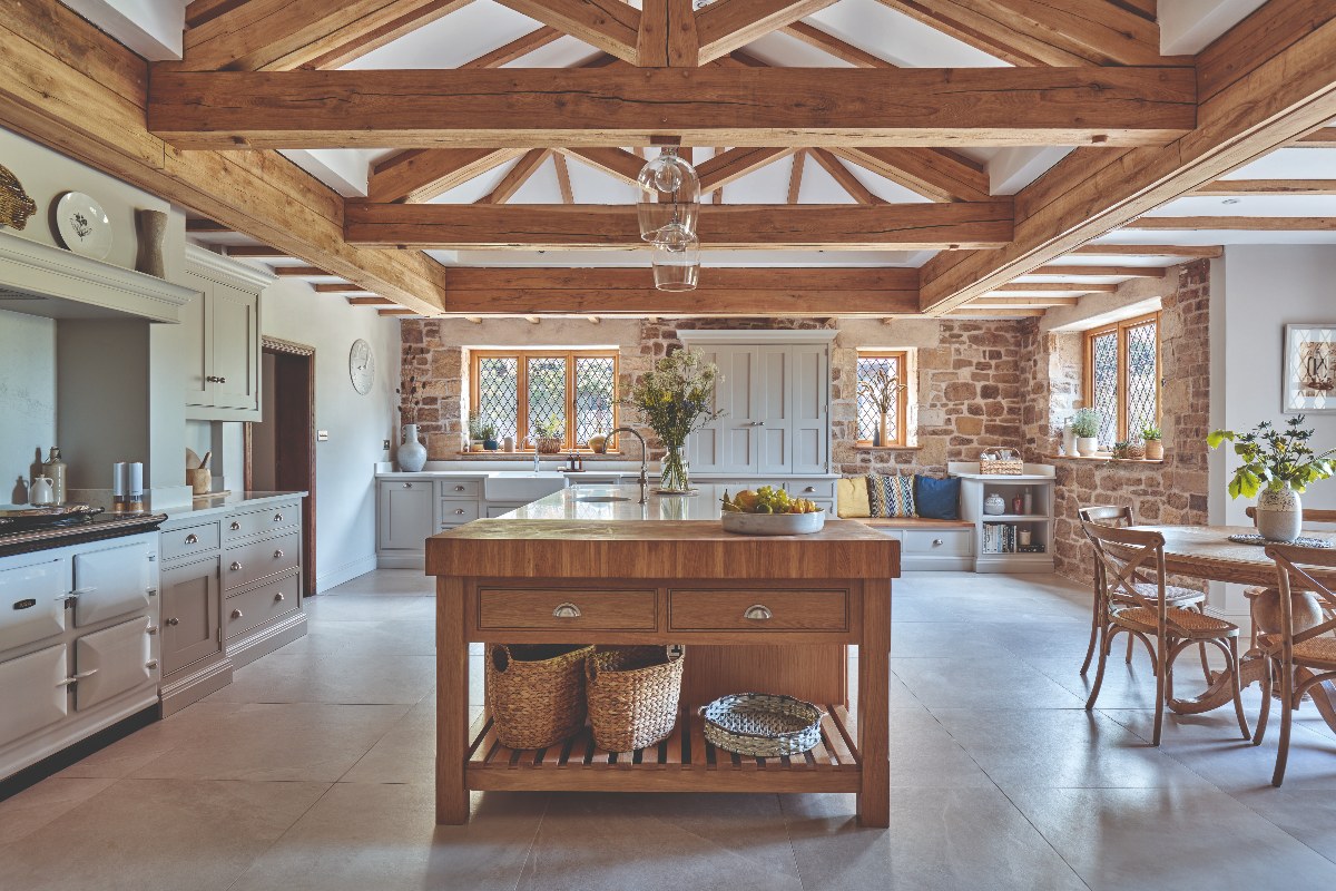 A country style kitchen with wood beams, classic cabinetry, a central island with a wood work table at one end and a compact dining area next to it.
