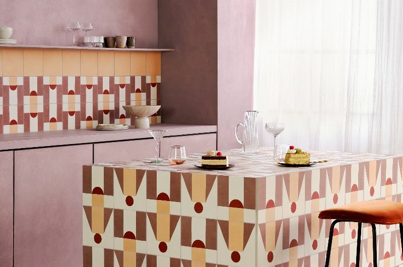 Kitchen design with pink handleless units and a patterned tile splashback, with a central island in matching tiles.