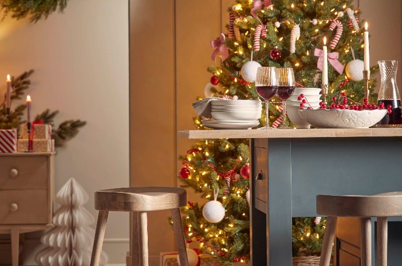 A farmhouse style kitchen with a freestanding island with weathered oak stools, and festive decorations, with a tree in the background.
