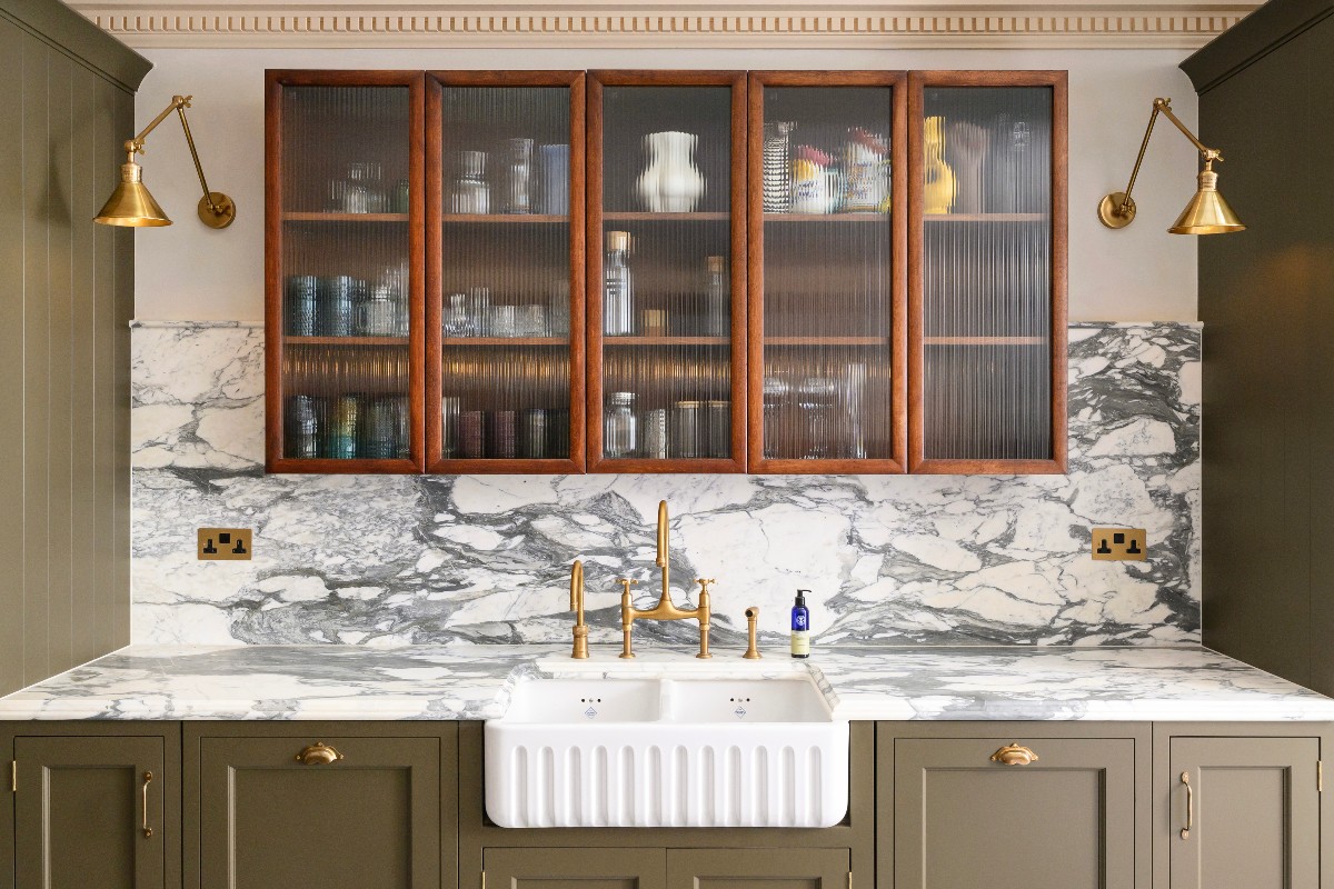 A sink zone with a fluted double butler bowl, green cabinetry, veiny marble, brass accents and a wall-mounter wood dresser with fluted glass.