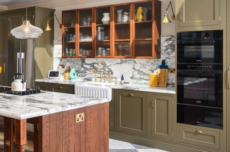 A farmhouse kitchen with olive green cabinets, built-in appliances, marble surfaces, checkerboard flooring and a central wood island with fluted glass cabinets.