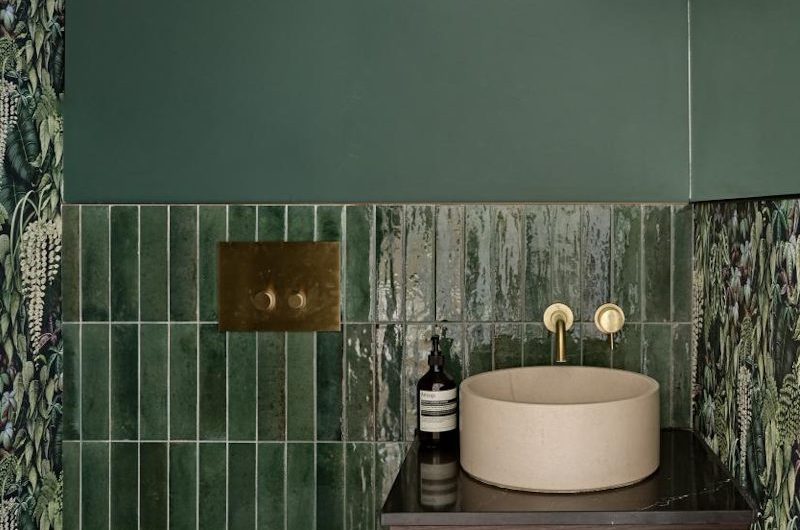 A cloakroom design with green metro tiles, dark green paint and moody wallpaper on the walls, a wood wall-mounted vanity with a countertop basin and a white wall-hung toilet. 