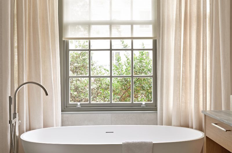 Neutral bathroom design with a freestanding bath, cream curtains, elegant decor and a wide window bringing in light.