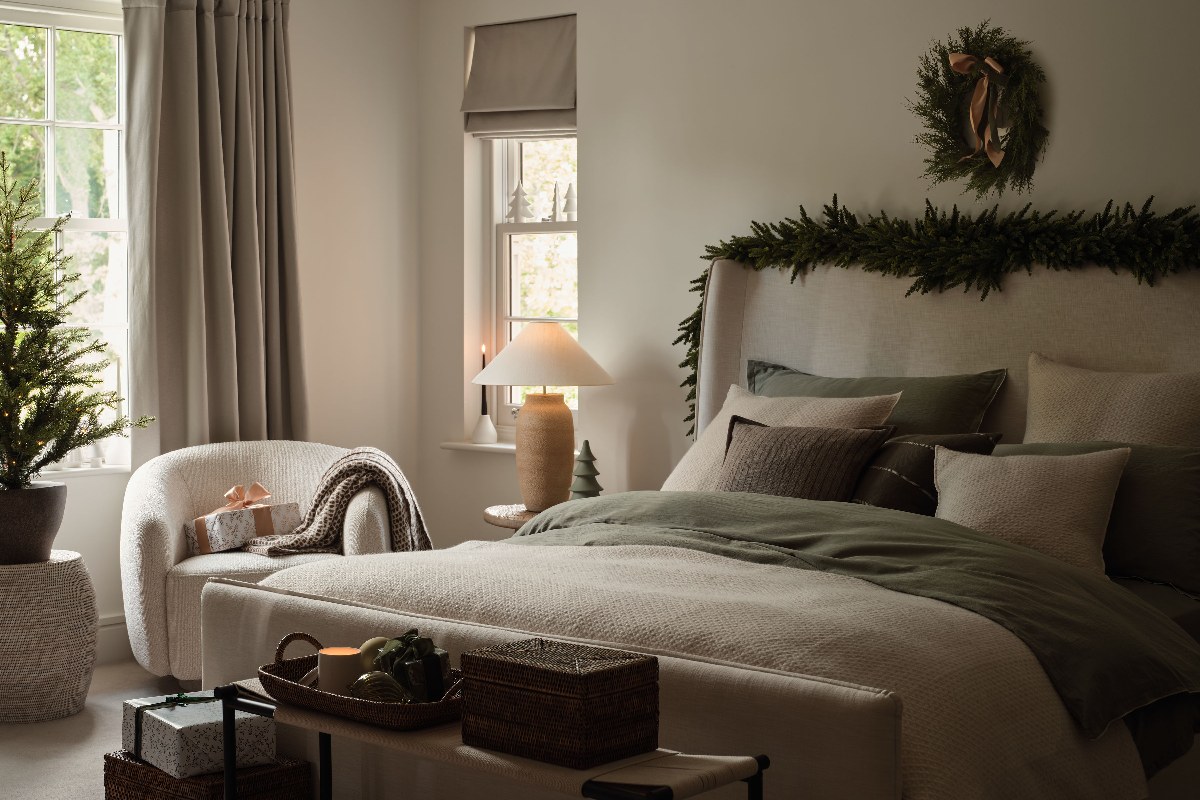 Neutral bedroom with potted trees, green garlands, neutral linen, champagne-coloured furnishings, and sleek candlesticks