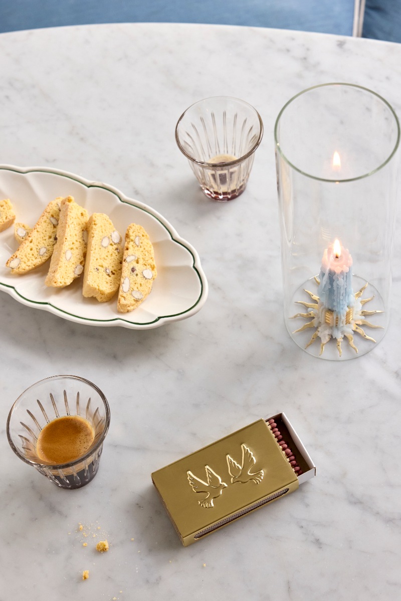 A marble table with a quirky plate with biscotti, a candle, a cup of coffee and a brass matchbox sleeve.
