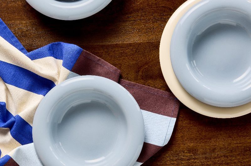 A wood table with blue and cream pasta bowls and patterned tea towels.
